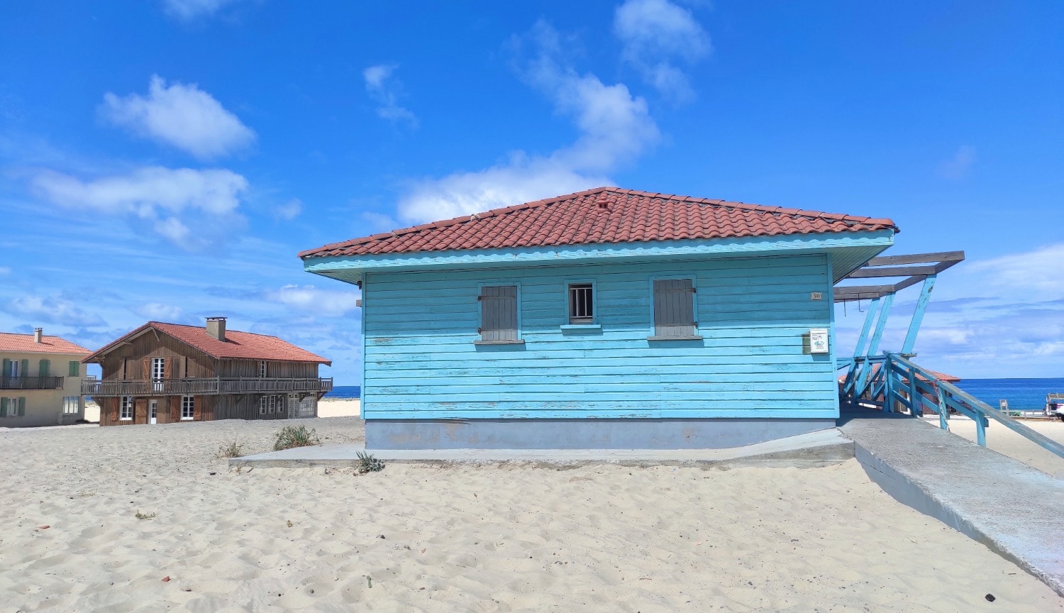Cabane en bois bleue à Vielle-Saint-Girons sur la plage des Landes, avec vue sur l’océan Atlantique et maisons typiques en arrière-plan.